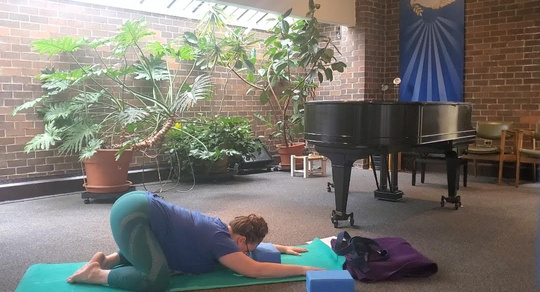 A fat white woman in a blue shirt and teal mesh leggings is demonstrating child's pose or balasana using a yoga block underneath her forehead. In the background are many large green leafy plants, a stool, a grand piano, and other yoga props