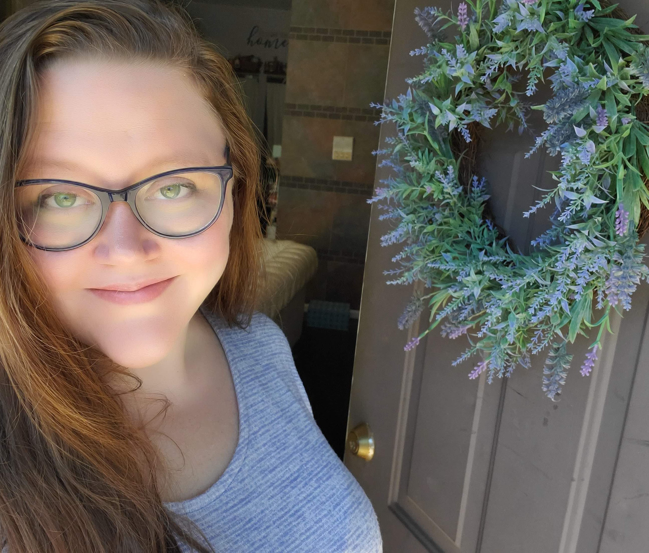 Yoga teacher with long brown hair, white skin, black cat eye glasses, and blue sweater stands casually in a doorway with a lavender wreath as though to welcome you in.